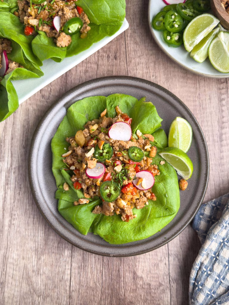 Asian turkey lettuce wraps on a rustic plate, topped with sliced jalapeños, radishes, and chopped peanuts, served with lime wedges on a wooden table. A fresh, healthy, and gluten-free meal option.