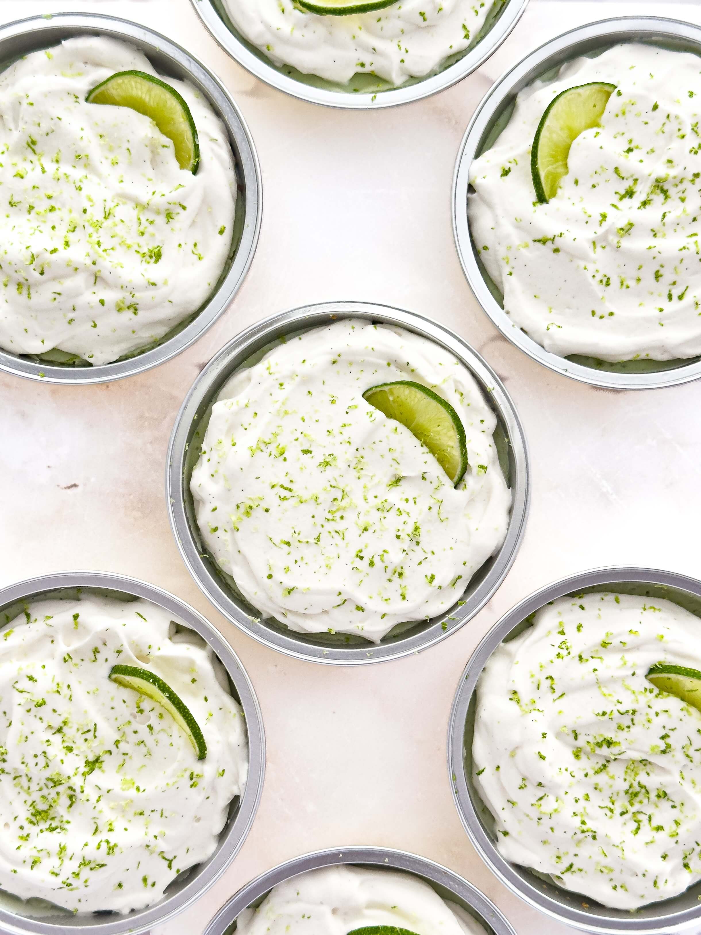 Gluten-free mini key lime pies topped with whipped cream, fresh lime slices, and lime zest, served in individual metal tins on a light background