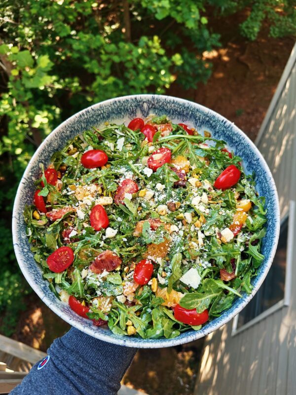 Overhead view of a fresh gluten-free arugula citrus salad with blood oranges, cherry tomatoes, pistachios, and shaved Grana Padano cheese served in a blue ceramic bowl outdoors.