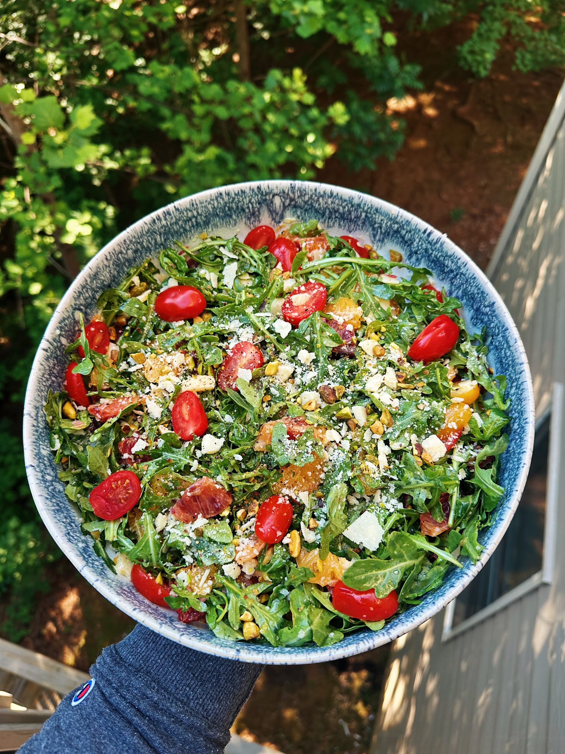 Overhead view of a fresh gluten-free arugula citrus salad with blood oranges, cherry tomatoes, pistachios, and shaved Grana Padano cheese served in a blue ceramic bowl outdoors.